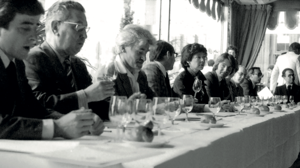Group of people sitting at a long table with glasses and cups, possibly in a restaurant or meeting setting. The Judgement of Paris