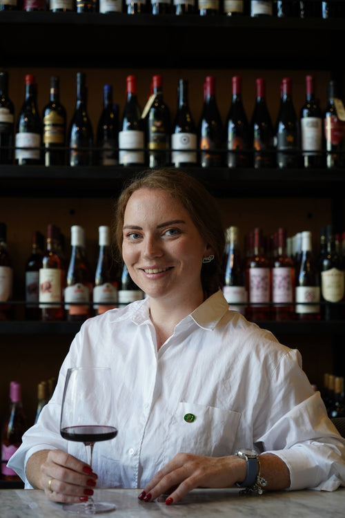 Daria Kholodilina sits at a desk behind a wall of wine bottles, holds a glass of red wine with her right hand and smiles at the camera