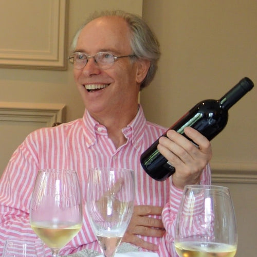 David is laughing and holding a bottle of wine, looking away from the camera. There are three wine glasses assembled in front of him.