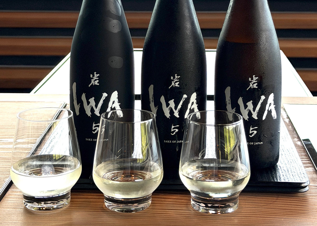 Three IWA 5 sake bottles on a wooden table, with three glasses in front of them