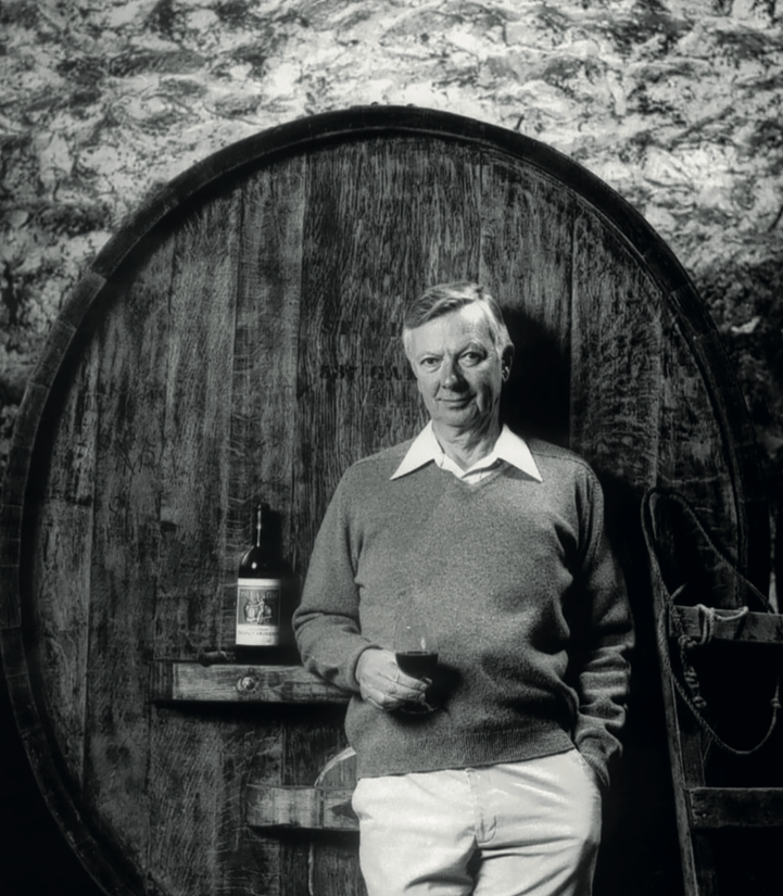 Man standing next to a large wooden barrel holding a bottle of whiskey.