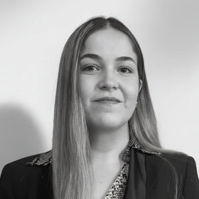 Lana-Nadia Koleilat with long light brown hair wearing a black blazer over a snakeskin-patterned top, standing against a plain light gray wall.