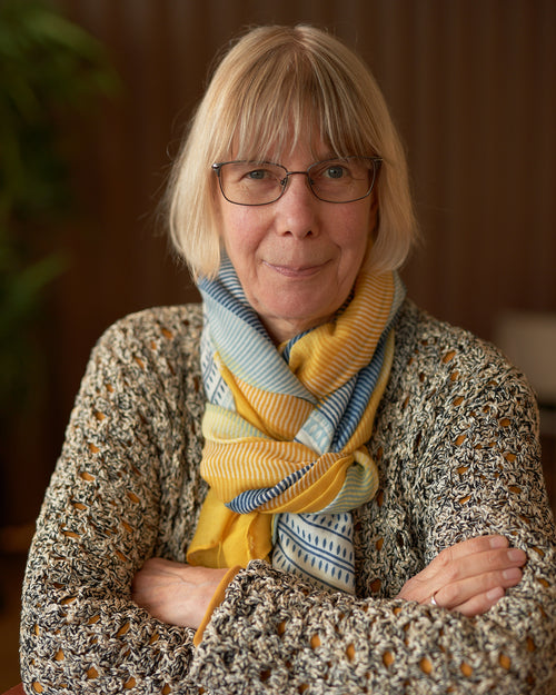 Wink Lorch stands looking at the camera with folded arms against a brown wall with a plant on her right